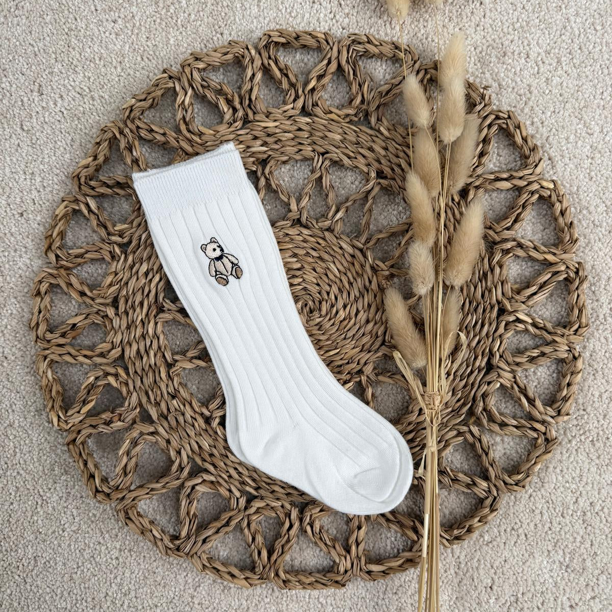 A single white ribbed crew sock with a small embroidered bear graphic on the cuff, laid flat on a round woven straw mat with dried bunny tail grass stems, on a beige carpet background.
