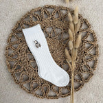 A single white ribbed crew sock with a small embroidered bear graphic on the cuff, laid flat on a round woven straw mat with dried bunny tail grass stems, on a beige carpet background.